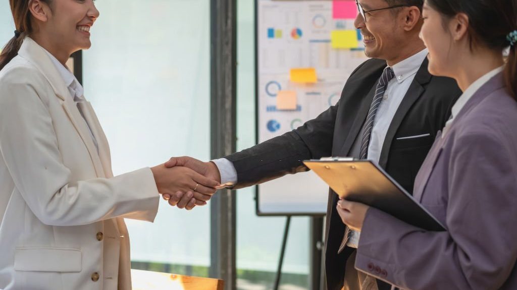 Business professionals shaking hands in a modern office setting, symbolizing customer acquisition and collaboration, with market research charts in the background.